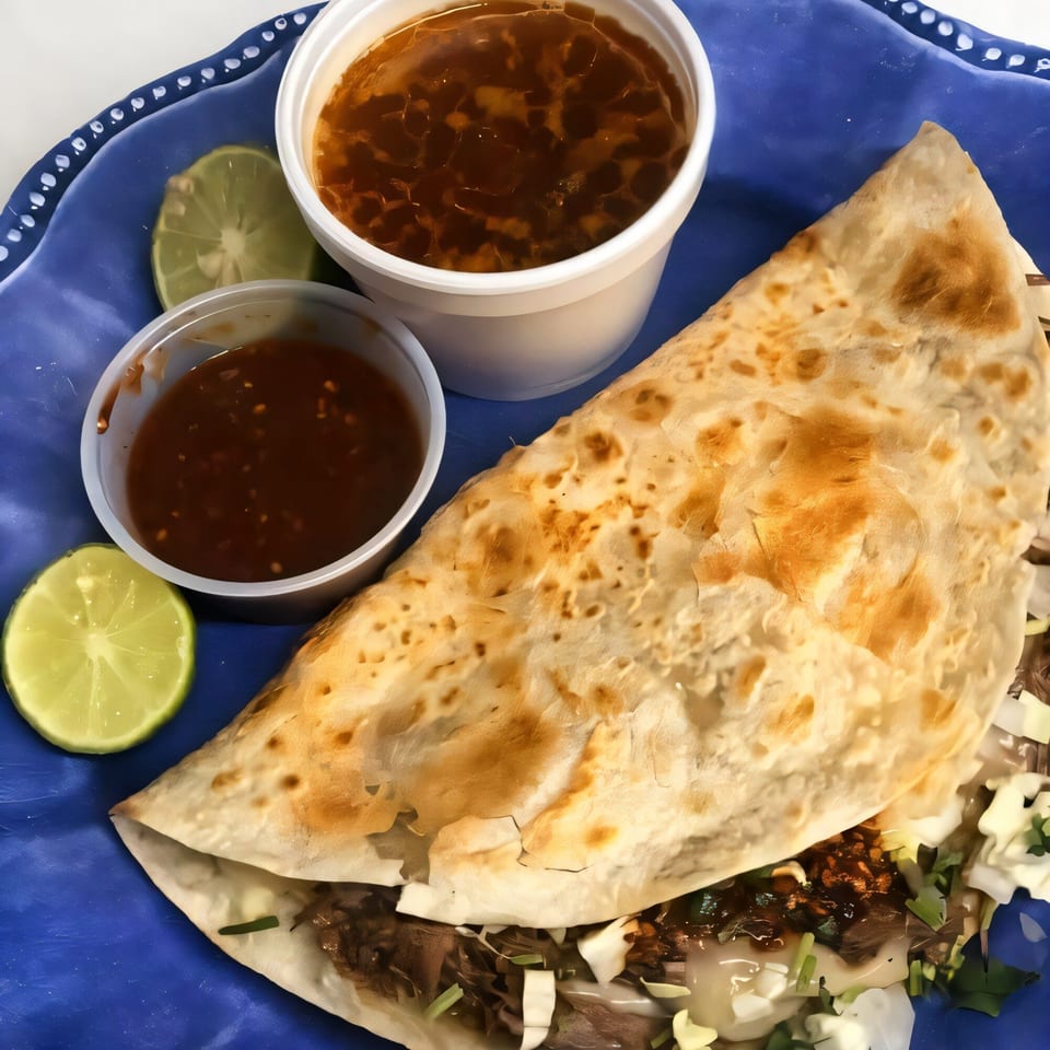 A large quesadilla with meat, cabbage, and sauce, served with consome and salsa on a blue plate.