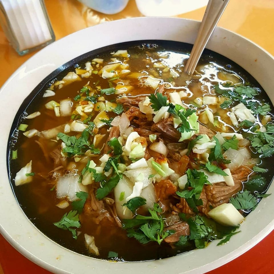 A close-up of a bowl of Birrieria Del Pueblo's birria, a rich Mexican stew with meat, onions, and cilantro.