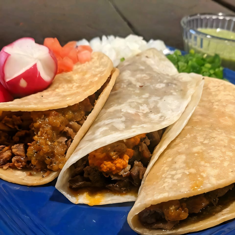 Three Birrieria Del Pueblo tacos on a blue plate with radish, tomato, and green sauce.